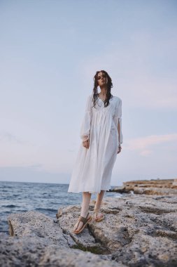 Beyaz elbiseli kadın taştan kumsal okyanus manzarasında duruyor.