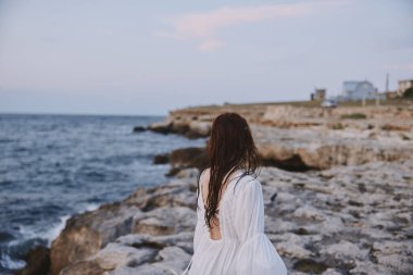 Beyaz elbiseli kadın plaj taşlarında yürüyor.