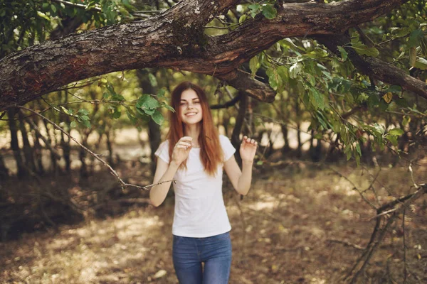 Tişörtlü ve kotlu bir kadın parkta yeşil yapraklı bir ağacın yanında duruyor. — Stok fotoğraf