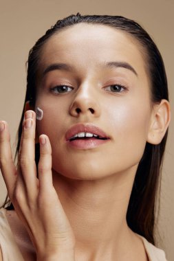 Genç bir kadın rahatlatıcı bir yüz maskesi takıyor. Kozmetik bej arka plan.