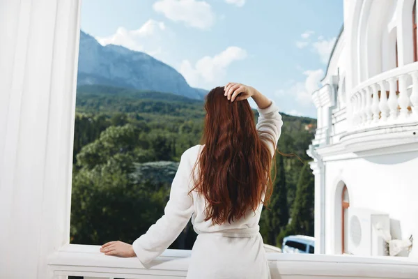 Una mujer con una túnica blanca admira la vista de las montañas desde el balcón sosteniendo su cabello — Foto de Stock