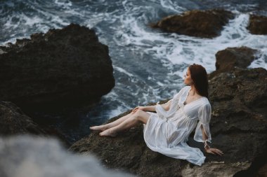 Islak saçlı, beyaz elbiseli, çıplak ayaklı bir kadın uçurumun kenarında oturuyor.