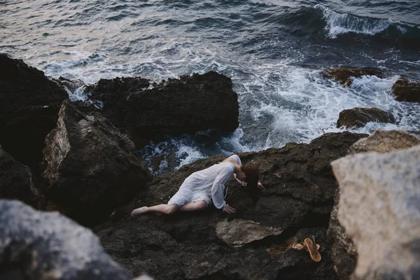 Barefoot woman in long white dress wet hair lying on a rocky cliff view from above — Stock Photo, Image