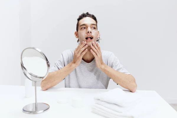 Chlap s kudrnatými vlasy v bílém tričku sedí u stolu se zrcadlem a ručníkem krém na obličeji zdraví — Stock fotografie