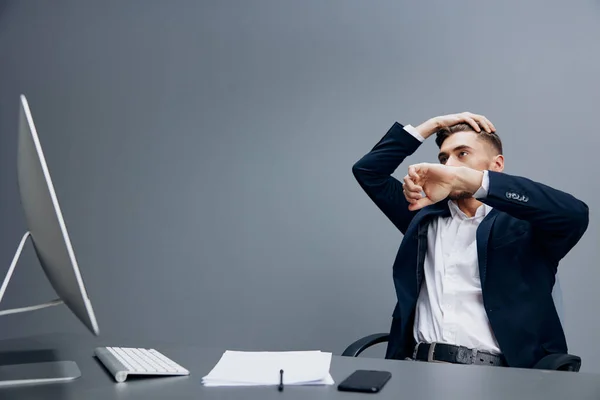 Egy öltönyös férfi telefonos érzelmekkel dolgozik egy számítógép előtt szürke háttér — Stock Fotó