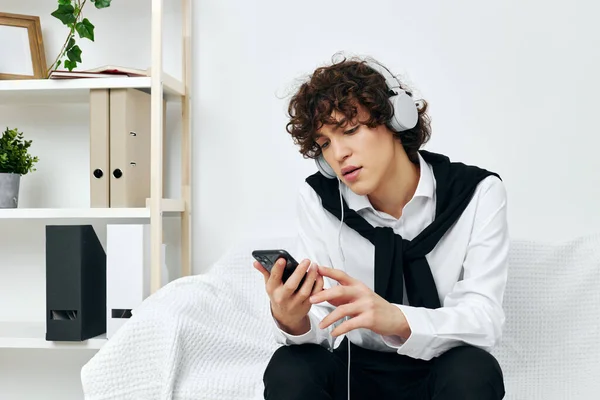 Tipo con el pelo rizado se sienta en el sofá con auriculares en el teléfono —  Fotos de Stock