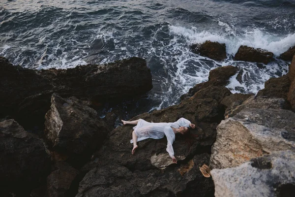 長い白いドレスを着た女が岩だらけの崖の風景の上に横たわっている — ストック写真