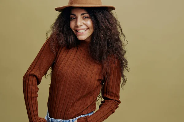 Enjoyed stylish curly Latin female in brown hat, smiling at camera, holding hand in jeans, saying Yeah, isolated green background. Copy space clothing fashion brands, free place for your ad. — Stock Photo, Image