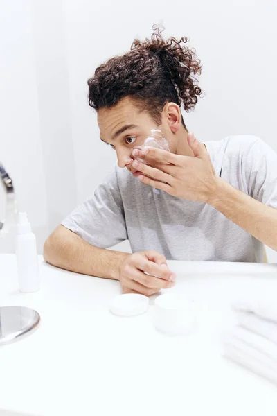 Guy with curly hair puts cream on the face but sitting at the table in front of the mirror isolated background — Stock Photo, Image