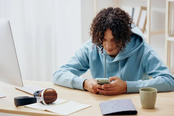 Mavi kazaklı kıvırcık saçlı genç bir adam telefonlu bir masada oturuyor. — Stok fotoğraf