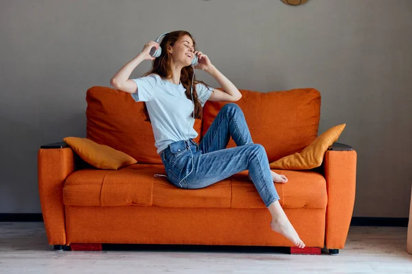 Sorrindo mulher ouvindo música com fones de ouvido no sofá laranja apartamentos — Fotografia de Stock