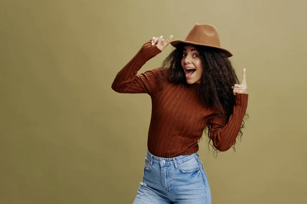 Enjoyed stylish curly Latin female in brown hat, smiling at camera, holding hand on hat, pointing finger up, isolated green background. Copy space clothing fashion brands, free place for your ad. — Stock Photo, Image