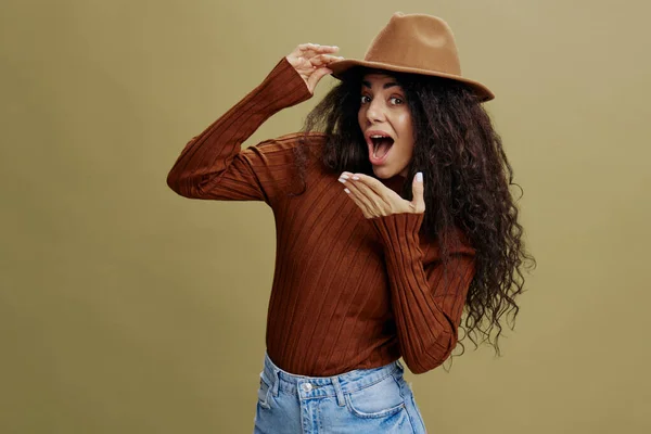 Wauw idee. Genieten van stijlvolle krullende Latijnse vrouw met bruine hoed, glimlachen op de camera, hand op de hoed, het sluiten van de mond, geïsoleerde groene achtergrond. Kopieer ruimte kleding modemerken, gratis plek voor uw advertentie. — Stockfoto