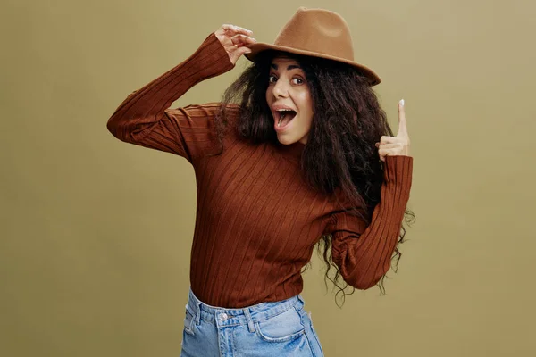 Great Idea concept. Overjoyed stylish curly Latin woman in brown hat, smiling at camera, holding hand on hat, pointing up, isolated green wall. People emotions lifestyle concept. Mock up copy space. — Stock Photo, Image