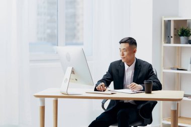 Takım elbiseli bir kahve bardağının içindeki yönetici bilgisayarın önündeki masada oturuyor.