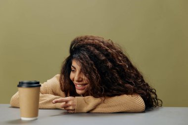 Gülümseyen mutlu, bronzlaşmış kıvırcık Latin kadın kahve fincanına bakar, uzanır zeytin yeşili arka planda izole bir şekilde masaya oturur. Uzayı kopyala Mockup Banner. Kahve sever misin?