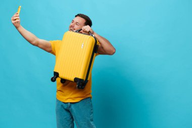 Sarı tişörtlü neşeli, bronzlaşmış yakışıklı adam mavi stüdyo arka planında izole edilmiş bir şekilde duran telefon duruşu bavuluyla selfie görüntülü görüşme yapıyor. Uzayın afiş modelini kopyala. Seyahat konsepti