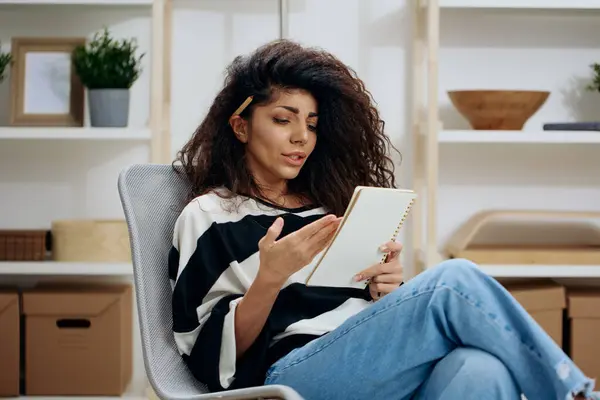 Concentrated pensive pretty beautiful curly tanned Latin woman with pen in hair in striped shirt hold notebook look down read work letter essay in home interior. Copy space Banner. Writer concept