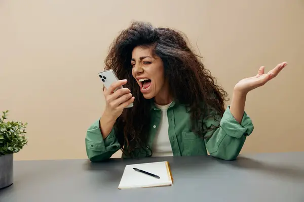 Angry crazy irritated tanned curly Latin lady scream at phone screen sit at the table isolated over pastel beige background. Copy space Mockup Banner. Concept call from debt collectors