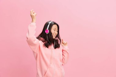 Pembe kapüşonlu, sevimli kulaklıklı Asyalı genç bayan pembe stüdyo arka planında izole edilmiş şekilde müzik eşliğinde dans ediyor. İyi bir teklif. Ses yayını platform reklam kavramı