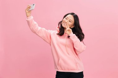 Pembe kapüşonlu tişörtlü neşeli gülümseyen Asyalı genç bayan pembe stüdyo arka planında izole edilmiş bir şekilde selfie görüntülü görüşme mesafesinde. İyi bir teklif. Gadget bağımlılığı Sosyal Medya kavramı