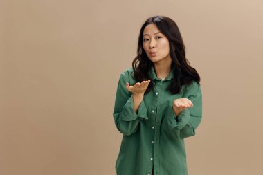 Haki yeşil tişörtlü mutlu Asyalı genç bir kıza aşık olmak, bej renkli pastel stüdyo arka planında izole bir şekilde poz vererek kameraya öpücük yolluyor. Güzel moda teklifi. Yaşam tarzı ve Duygular kavramı