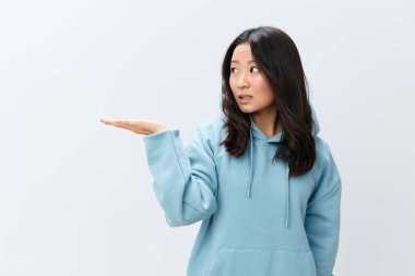 Kafası karışmış, mavi kapüşonlu genç Asyalı kadın beyaz stüdyo arka planında yalnız poz vererek avuç içini kaldırıyor. Reklam için en iyi teklif. Moda Yeni Koleksiyon Reklam Konsepti