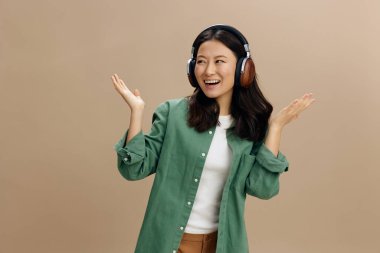 Hey buraya bak. Haki yeşili kulaklıklı neşeli mutlu Asyalı genç kadın bej renkli pastel stüdyo arka planında izole edilmiş şekilde avuç içlerini kaldırıyor. Güzel moda teklifi. Müzik Uygulaması Platformu Reklam Konsepti