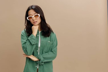 Haki yeşil tişörtlü, üzgün Koreli genç kadın bej renkli pastel stüdyo arka planında izole edilmiş yumruk pozuna yaslanıyor. Güzel moda teklifi. Güneş gözlüğü reklam konsepti