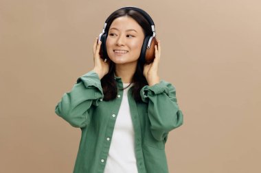 Afiyet olsun. Hakiki yeşil tişörtlü mutlu Asyalı genç bayan kulaklıklara dokunuyor bej renkli pastel stüdyo arka planında yalnız poz veriyor. Güzel moda teklifi. Müzik Uygulaması Platformu Reklam Konsepti