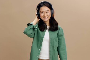 Bu sesin tadını çıkar. Hakiki yeşil tişörtlü mutlu Asyalı genç bayan bej renkli pastel stüdyo arka planında izole edilmiş şekilde duran kulaklıklara dokunuyor. Güzel moda teklifi. Müzik Uygulaması Platformu Reklam Konsepti