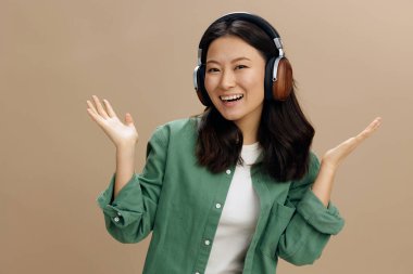 Hey buraya bak. Haki yeşili kulaklıklı neşeli mutlu Asyalı genç kadın bej renkli pastel stüdyo arka planında izole edilmiş şekilde avuç içlerini kaldırıyor. Güzel moda teklifi. Müzik Uygulaması Platformu Reklam Konsepti