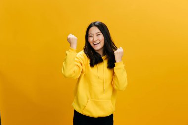 Evet, başardım. Aşırı neşeli Asyalı öğrenci, sarı kapüşonlu genç kadın. Yumruklarını kaldır ve gülümse. Turuncu stüdyo arka planında izole bir şekilde poz ver. Güzel teklif. Halkın Duyguları kavramı