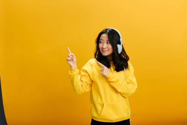 Vay canına, şuna bak. Sarı kapüşonlu kapüşonlu, sevimli Asyalı genç bayan parmak işareti dışında turuncu stüdyo arka planında yalnız poz veriyor. Güzel teklif. Mükemmel çalma listesi Ses Uygulaması kavramı
