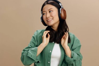 Afiyet olsun. Hakiki yeşil tişörtlü mutlu Asyalı genç bayan kulaklıklara dokunuyor bej renkli pastel stüdyo arka planında yalnız poz veriyor. Güzel moda teklifi. Müzik Uygulaması Platformu Reklam Konsepti