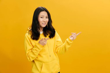 Sarı kapüşonlu kapüşonlu genç Asyalı kafası karışık genç bayan turuncu stüdyo arka planında yalnız poz vermek dışında fotokopi alanı gösteriyor. Güzel teklif. Halkın Duyguları kavramı