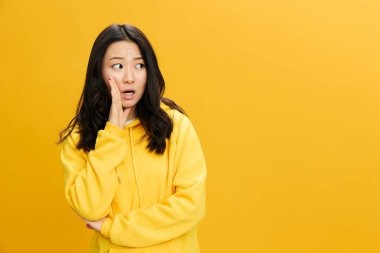 Sarı kapüşonlu tişörtlü, heyecanlı Asyalı genç bayan ağız kenarında el ele tutuşurken turuncu stüdyo arka planında gizli pozlar veriyor. Güzel teklif. Halkın Duyguları kavramı