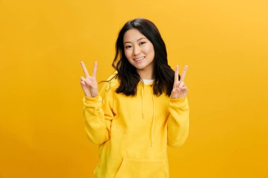 Keyfin yerinde. Sarı kapüşonlu tişörtlü neşeli Asyalı genç bayan turuncu stüdyo arka planında izole edilmiş V işareti gösteriyor. Güzel teklif. Halkın Duyguları konsepti. Boşluğu kopyala