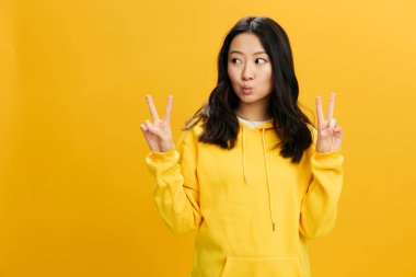 Keyfin yerinde. Sarı kapüşonlu tişörtlü neşeli Asyalı genç bayan turuncu stüdyo arka planında izole edilmiş V işareti gösteriyor. Güzel teklif. Halkın Duyguları konsepti. Boşluğu kopyala