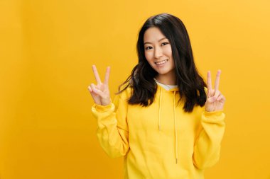 Keyfin yerinde. Sarı kapüşonlu tişörtlü neşeli Asyalı genç bayan turuncu stüdyo arka planında izole edilmiş V işareti gösteriyor. Güzel teklif. Halkın Duyguları konsepti. Boşluğu kopyala
