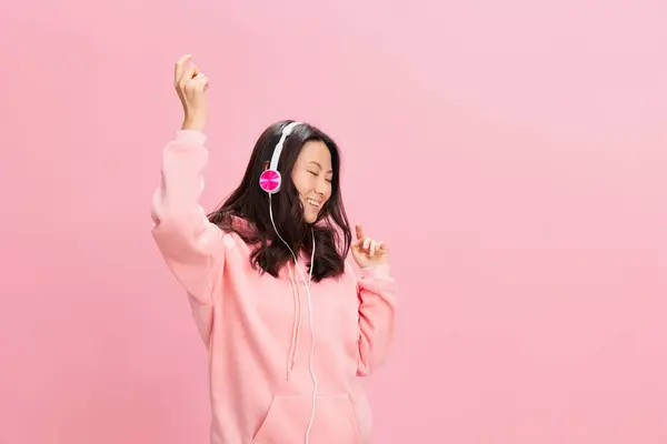 ピンクのフードージーのスウェットシャツで ピンクのスタジオの背景に隔離された音楽に移動するかわいいヘッドフォンダンスでアジアの学生の若い女性をお楽しみください 良いオファーだ サウンドストリーミングプラットフォーム広告コンセプト — ストック写真