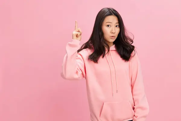 看这里 身穿粉色连帽衫的兴奋的亚洲学生年轻女士用手指抬起头 在粉红的工作室背景上摆出孤立的姿势 很好的提议时装新系列概念 — 图库照片