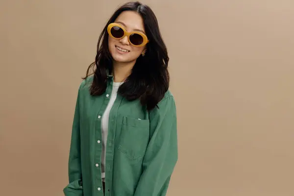 Cheerful happy lovely Korean young woman in khaki green shirt stylish eyewear smiles at camera posing isolated on over beige pastel studio background. Cool fashion offer. Sunglasses ad concept