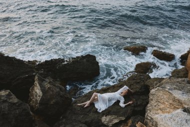 Uzun beyaz elbiseli, çekici bir kadın yukarıdan kayalık bir uçurumun kenarında ıslak saçlarıyla yatıyor. Yüksek kalite fotoğraf