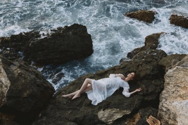 Uzun beyaz elbiseli, çıplak ayaklı, ıslak saçlı bir kadın yukarıdan kayalık bir uçurumun kenarında yatıyor. Yüksek kalite fotoğraf