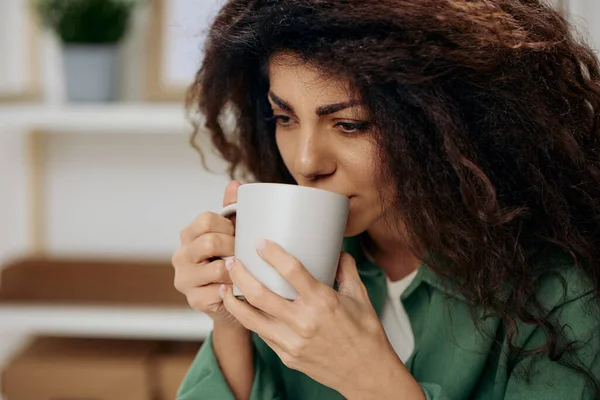 Frustrated sad tanned curly Latin lady troubled with problem with cup of tea coffee in casual shirt sit on chair in home modern interior background look aside. Copy space Banner. Concept psychology