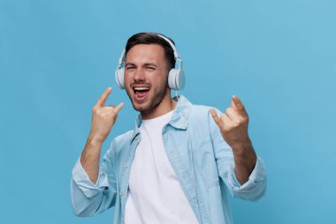 Sıradan tişört kulaklıklarıyla bronzlaşmış yakışıklı adam mavi stüdyo arka planında izole edilmiş korna rock hareketinin favori şarkı gösterisini dinle. Uzayın afiş modelini kopyala. Müzik konsepti