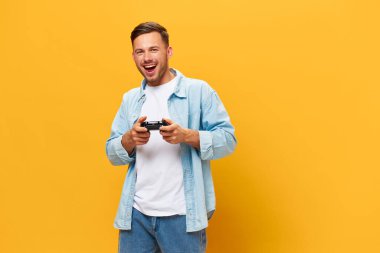 Mavi tişörtlü, bronz tenli yakışıklı bir adam turuncu sarı stüdyo arka planında izole edilmiş joystick pozu veriyor. Uzayın afiş modelini kopyala. İnsanların yaşam tarzı Şanslı kazanan konsepti