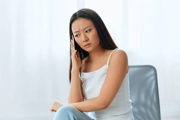 Wary confused attended tanned lovely young Asian woman listen shocked news at call using phone at home interior living room. Distance communication Social media concept. Cool offer Banner Wide angle