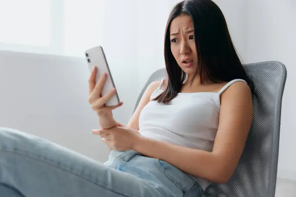 Irritated shocked young Asian woman read fake bad news in messages sit on chair at home interior living room. Distance communication, Remote work, Social media concept. Cool offer Banner Mockup
